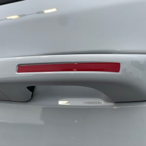 May include: White car door handle with a red accent strip. The handle is made of plastic and has a smooth surface.