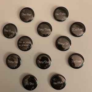 May include: A set of 12 black button badges with white text and star designs. The badges feature various pronoun combinations, including She/Her, He/They, They/Them, They/It, They/He, They/She, It/Its, He/Him, Xe/Xem, and a custom option.