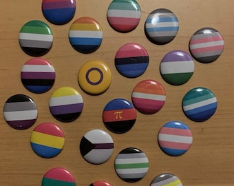 Op de afbeelding: Een verzameling van 18 ronde knopen met verschillende pride-vlaggen, waaronder de transgender-, lesbische-, homoseksuele-, biseksuele-, panseksuele-, aseksuele-, non-binaire- en interseksevlaggen. Er is ook een knoop met een gele cirkel en een zwart symbool, en een andere knoop met een zwarte achtergrond en het symbool "π".