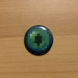 Puede incluir: Un botón de ojo pixelado verde y negro. El ojo está rodeado por un borde azul oscuro.