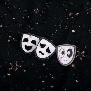 May include: Three white theatre masks with different expressions: happy, sad, and neutral. The masks are set against a black background with gold star-shaped glitter.