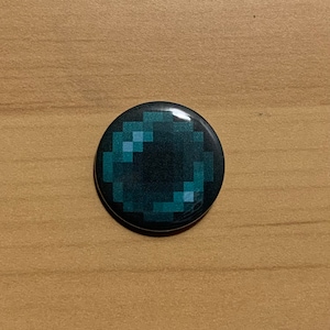 Puede incluir: Un botón redondo con una imagen pixelada de una perla de Ender verde oscuro y azul del videojuego Minecraft.