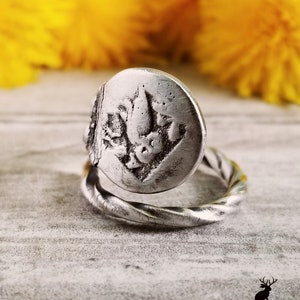 May include: A silver ring with a twisted band and a round, textured top featuring a floral design.