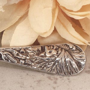 May include: A silver spoon with an ornate handle and a detailed bowl. The bowl features an engraved image of a building and the text "FURNITURE FACTORY" and "GRAND RAPIDS MICH". The handle has a floral design.