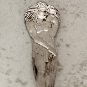 May include: A silver spoon with a detailed Native American figure on the handle. The figure is sculpted with intricate details, and the spoon has a decorative floral design on the stem. The spoon is marked with the text "SISTER DANDY".