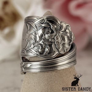 Puede incluir: Un anillo de plata hecho con una cuchara, con un diseño floral. El anillo presenta un patrón floral en relieve detallado en la parte delantera, con hojas y pétalos. La banda del anillo está hecha del mango de la cuchara, con líneas grabadas.
