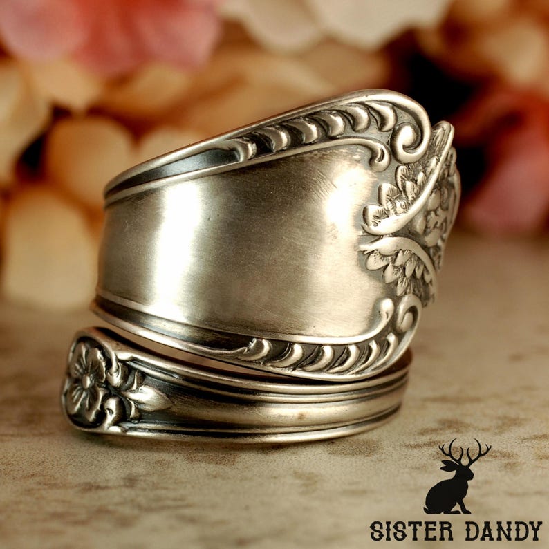 Puede incluir: Un anillo de plata hecho a mano a partir de una cuchara, con intrincados detalles florales y de volutas. El anillo tiene un dise&ntilde;o &uacute;nico y envolvente, que muestra la forma original de la cuberter&iacute;a reutilizada. Una pieza de joyer&iacute;a llamativa.
