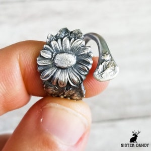 Puede incluir: Un anillo de plata con un diseño de margarita. El anillo está hecho de una pieza de metal retorcido y presenta una margarita detallada con hojas.