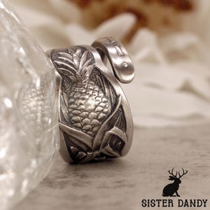 Puede incluir: Un anillo plateado con un diseño detallado de piña. El anillo presenta una superficie texturizada y una banda curva. La piña es el punto focal, con detalles intrincados de las hojas y la fruta. El anillo se muestra en primer plano, destacando la artesanía.