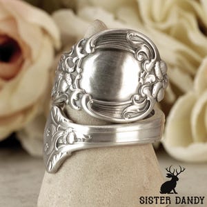 Op de afbeelding: Een zilverkleurige ring gemaakt van gerecycled bestek. De ring heeft sierlijke bloemen- en kruldetails rond een centraal ovaal ontwerp. De band is gevormd uit de steel van een lepel. De ring wordt gepresenteerd op een neutrale ondergrond.