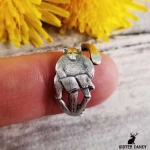 May include: Silver ring featuring a monkey holding a book. The monkey is sitting on a tree branch. The ring is on a finger.