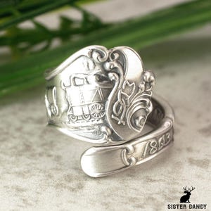 Puede incluir: Anillo de cuchara plateada con diseño de tren en relieve y el año 1843. El anillo presenta detalles ornamentados y una banda curvada, mostrando una estética vintage. Una pieza de joyería única.
