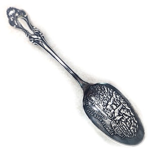 May include: A silver-toned souvenir spoon featuring an ornate, scrolled handle design. The bowl of the spoon displays a detailed embossed scene of a mountain landscape with the text SUMMIT OF PIKE'S PEAK ALTITUDE 14147 FEET engraved clearly.