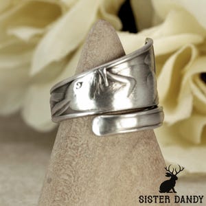 May include: A silver-toned wrap ring crafted from a repurposed spoon handle, featuring an embossed floral or leaf-like pattern along the band. The metal has a polished finish with visible indentations and curves characteristic of flatware.
