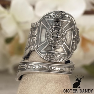 Puede incluir: Un anillo de cuchara plateado con un diseño de cruz en relieve dentro de un marco circular. El texto "LOOK UP" está inscrito alrededor de la cruz central. La banda muestra volutas decorativas y letras adicionales a lo largo del borde.