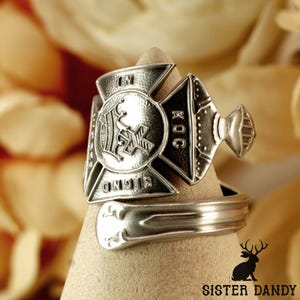 Könnte beinhalten: Ein silberner Löffelring mit einem detaillierten Emblem. Das Emblem zeigt Text und ein Kreuzdesign. Der Ring ist um eine beige Form gewickelt. Das Sister Dandy-Logo befindet sich unten rechts.