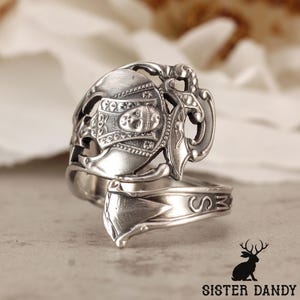 May include: A silver spoon ring with an ornate, detailed design. The ring features a decorative face and a band with the letters "SMZ". The ring is a unique piece of jewelry.
