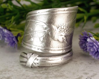 Puede incluir: Un anillo de cuchara plateado con un diseño detallado de colibrí y flores. El anillo se envuelve alrededor del dedo y presenta un acabado texturizado. Una pieza de joyería única.