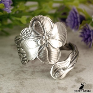Pode incluir: Um anel prateado com um design floral detalhado. O anel apresenta uma flor grande e aberta com detalhes intrincados nas pétalas. A banda curva-se para formar um ajuste ajustável. Uma peça de joalharia única.