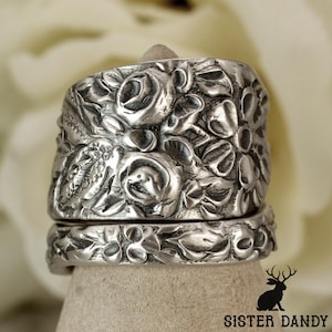 May include: Two silver rings with detailed floral designs, featuring roses and leaves. The rings are stacked on a neutral-colored cylindrical object, showcasing the intricate craftsmanship. The rings are likely for a wedding or engagement.