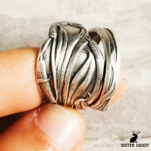 Puede incluir: Dos anillos de plata con diseños intrincados. El anillo superior presenta un patrón floral, mientras que el anillo inferior tiene un diseño más abstracto. Ambos anillos se llevan en un dedo.