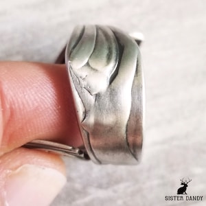 May include: A silver ring made from a repurposed spoon. The spoon has been hammered and shaped into a unique design. The ring is worn on a finger.