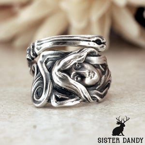 May include: A silver-toned metal ring featuring a sculpted relief of a face with flowing hair. The band wraps around in a bypass style, with oxidized detailing in the recessed areas to highlight the intricate facial features and hair texture.