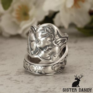 Puede incluir: Un anillo de plata hecho de una cuchara con un patrón floral en relieve en la parte superior ancha. La banda muestra letras grabadas que incluyen el texto parcial RCH y ET, con un acabado metálico pulido y áreas hundidas oxidadas.