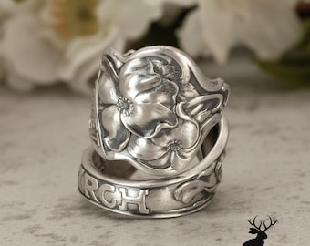 Puede incluir: Un anillo de plata hecho de una cuchara con un patrón floral en relieve en la parte superior ancha. La banda muestra letras grabadas que incluyen el texto parcial RCH y ET, con un acabado metálico pulido y áreas hundidas oxidadas.