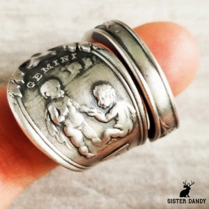 Könnte beinhalten: Ein silberner Ring mit dem Sternzeichen Zwillinge. Der Ring, vermutlich aus recyceltem Silberbesteck gefertigt, zeigt das Wort "GEMINI" über zwei cherubartigen Figuren. Der Ring wird an einem Finger getragen.