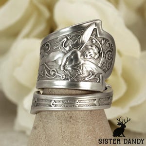 Puede incluir: Un anillo de metal plateado hecho a partir de una cuchara reutilizada, con un relieve de una figura alada rodeada de intrincados adornos y motivos florales. La banda muestra un patrón texturizado con formas geométricas repetitivas.
