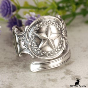 May include: A silver spoon ring with a raised star design in the center, surrounded by a laurel wreath. The ring has ornate detailing and a curved band. The ring is a unique piece of jewelry.