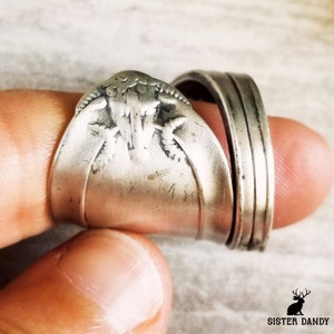 Può includere: Un anello a cucchiaio d'argento con un design a tre foglie. L'anello è indossato su un dito ed è impilato con una fascia d'argento.