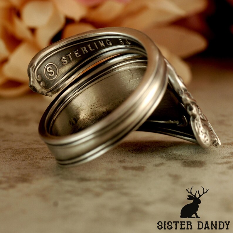 Puede incluir: Un anillo de plata hecho a mano a partir de una cuchara, con la palabra "STERLING" visible. El anillo tiene un dise&ntilde;o curvo &uacute;nico, que muestra la forma original de la cuchara. Una pieza de joyer&iacute;a.