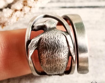 Puede incluir: Un anillo de plata con un diseño de pingüino detallado. El pingüino está en un marco circular elevado. El anillo se lleva en un dedo.