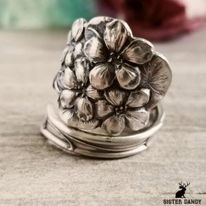 May include: A silver ring with a floral design. The flowers are made of metal and are arranged in a cluster. The ring is made of a thick band of metal.