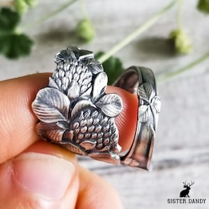 May include: A silver ring with a floral design. The ring features detailed leaves and a textured flower, with a band that appears to be crafted from a spoon. The ring is held up by a finger.