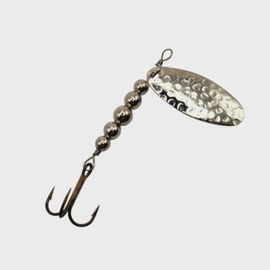 May include: A silver fishing lure with a hammered oval blade and a series of silver beads. The lure has a treble hook at the bottom. This fishing tackle is designed to attract fish.