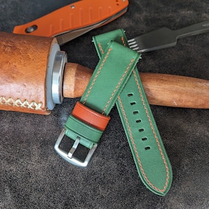 Può includere: Due cinturini per orologio in pelle verde con cuciture arancioni. Un cinturino ha una fibbia argentata. I cinturini sono su una superficie in pelle marrone.