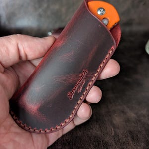 May include: A red leather knife sheath with a stitched edge and the text "Sohnwich" embossed on the front.