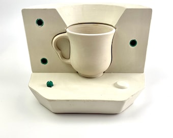 Puede incluir: Un molde de cerámica blanca para hacer una taza. El molde está hecho de yeso y tiene un acabado blanco liso. El molde está abierto por un lado y tiene un pequeño agujero por el otro lado. El molde está sentado sobre una superficie blanca.