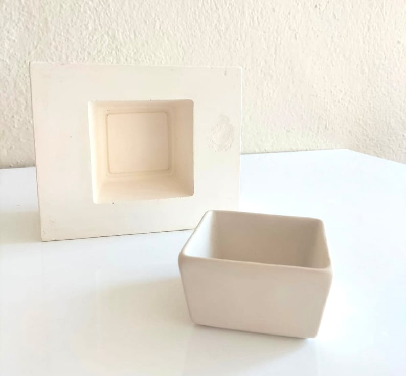 May include: A white ceramic mold for making a square planter, with a smaller square opening inside. A white ceramic square planter is shown next to the mold.