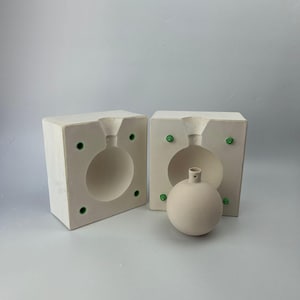 Puede incluir: Dos moldes de cerámica blanca para hacer adornos redondos. Los moldes están abiertos y un adorno de cerámica blanca está sentado en uno de los moldes.