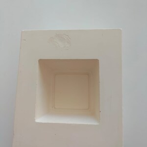 May include: White ceramic mold with a square opening in the center. The mold is designed for creating a square-shaped object.