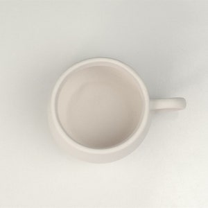 Pode incluir: Uma caneca de cer&acirc;mica branca com uma al&ccedil;a arredondada. A caneca est&aacute; vazia e tem um acabamento liso e fosco.