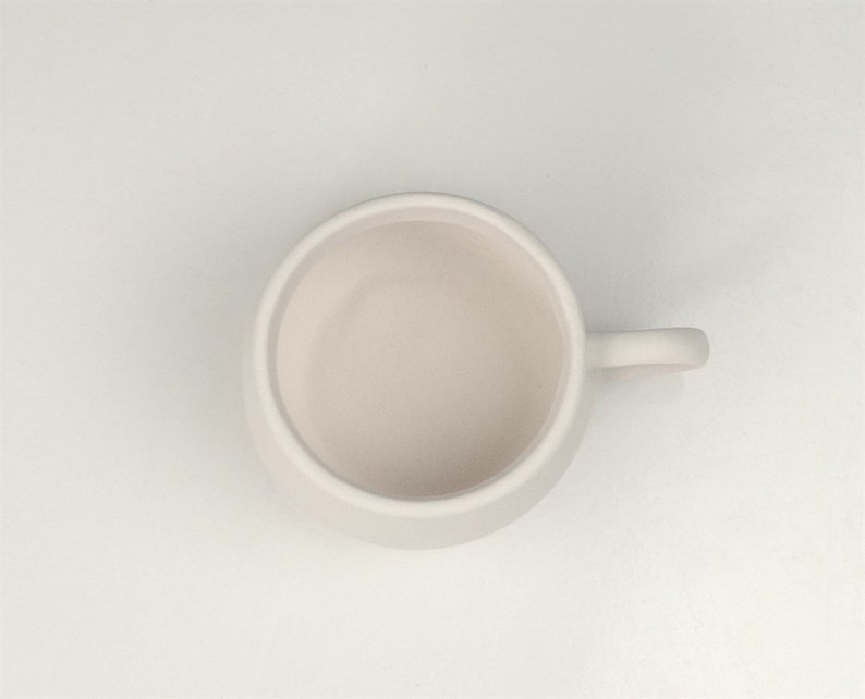 Pode incluir: Uma caneca de cer&acirc;mica branca com uma al&ccedil;a arredondada. A caneca est&aacute; vazia e tem um acabamento liso e fosco.