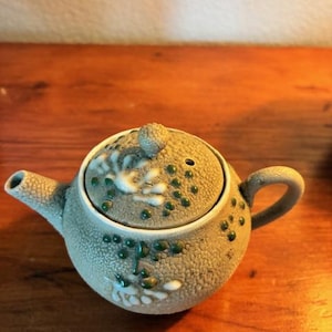 May include: A small, round, ceramic teapot with a textured, speckled surface. The lid and body are decorated with white and green floral designs. The teapot has a small spout and a curved handle.