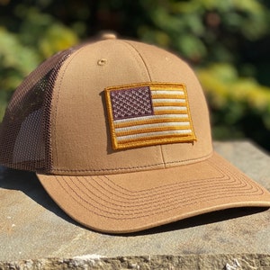 Peut inclure: Un chapeau de camionneur brun et beige avec un filet arrière brun. Le chapeau a un petit patch du drapeau américain sur le devant.