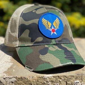 May include: Camouflage trucker hat with a blue and gold patch featuring a star and wings. The patch has the text "USAF" on it.
