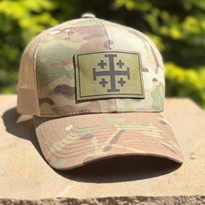 May include: A camouflage trucker hat featuring a rectangular olive green patch on the front panel. The patch displays a dark brown Jerusalem cross symbol. The hat has a structured front, a curved brim, and a tan mesh back for ventilation.